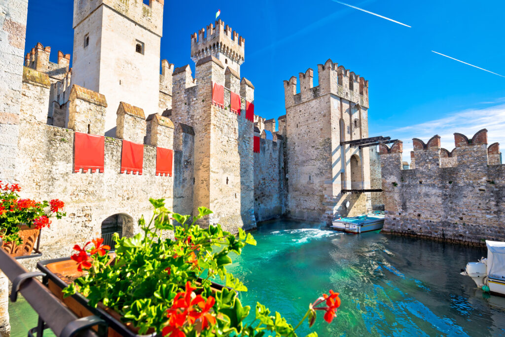 Castello medievale con mura e torri riflesse in un canale, circondato da fiori rossi.