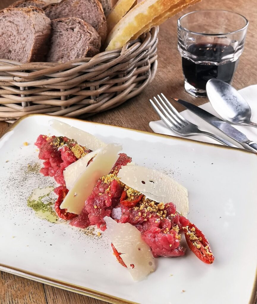 Tartare di carne con scaglie di formaggio e pane in un cesto sullo sfondo.