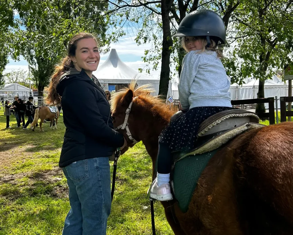 A woman and a young child with a helmet enjoy a pony ride in a sunny park.
