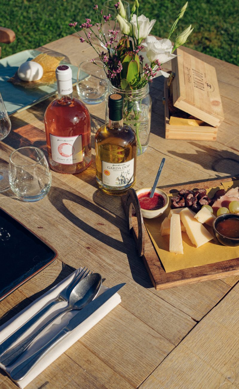 Tag alt e title suggeriti: "Tavolo da picnic con bottiglie di vino, formaggi, frutta secca e posate all'aperto."