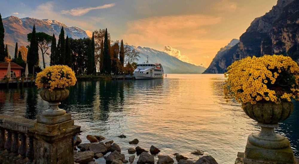 Tramonto su un lago alpino con montagne innevate e fiori gialli in primo piano.