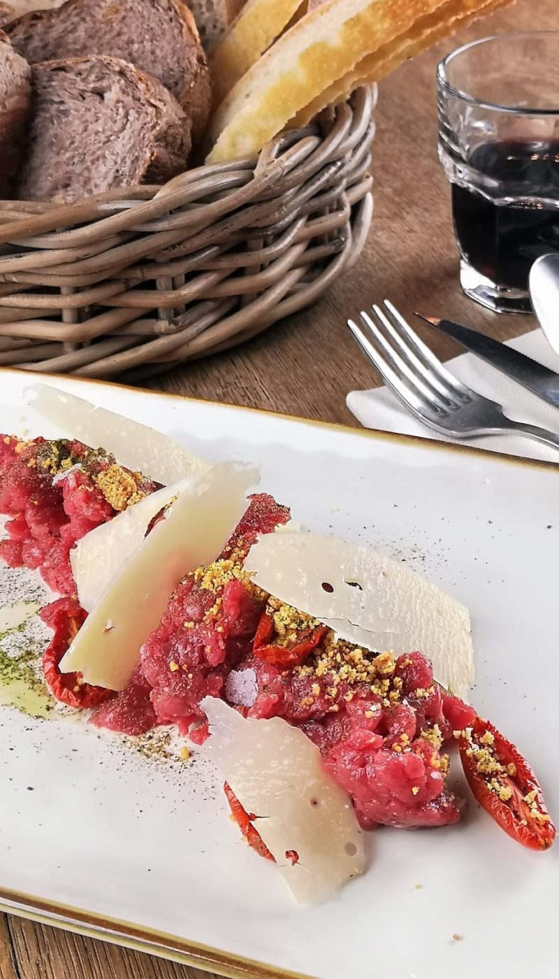 Tartare di carne con scaglie di formaggio e pane in un cesto sullo sfondo.