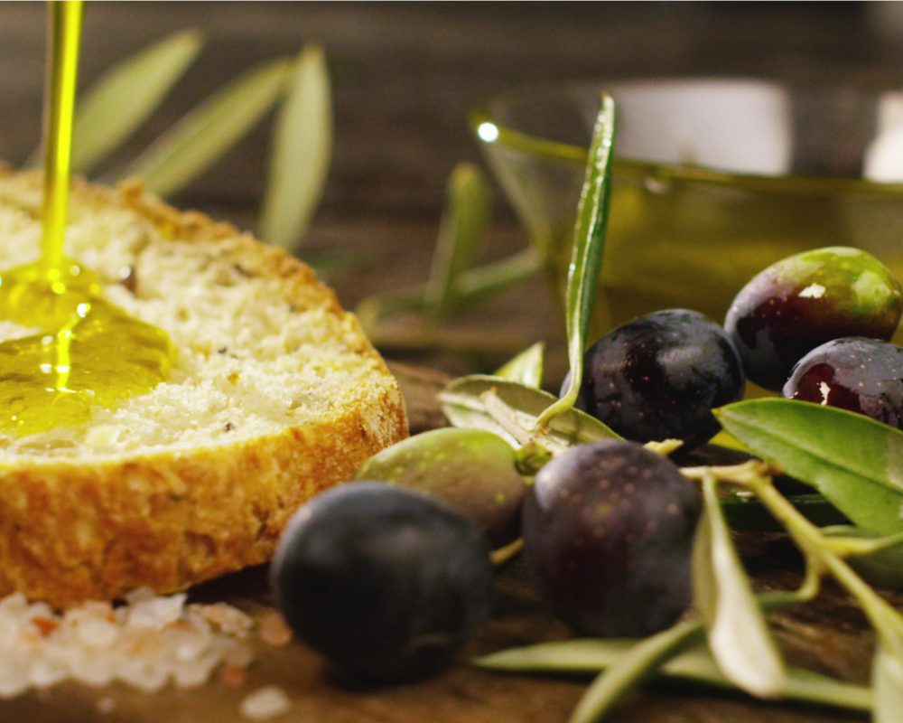 Fetta di pane con olio d'oliva versato sopra, circondata da olive e foglie.
